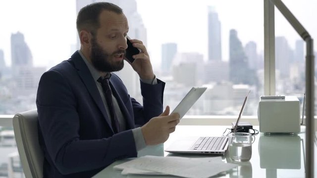 Angry businessman with tablet and laptop talking on cellphone in office
