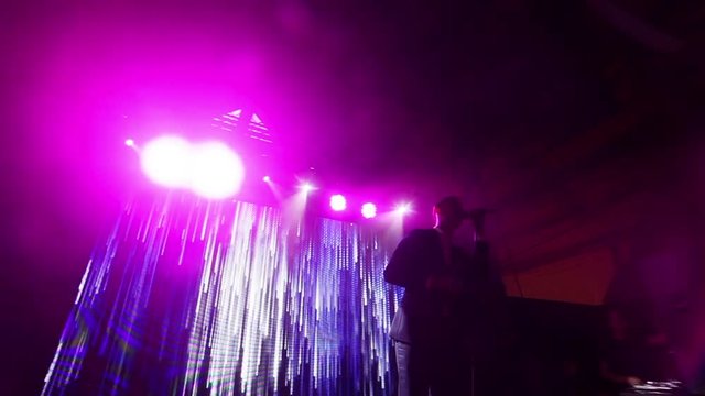 Silhouetted man sings on night club concert stage at LED screen background showing abstract animation of slowly moving waterfall lines POV. Stylish singer performs song with microphone at New Years