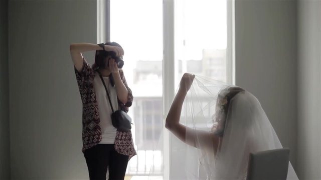 Photographer taking picture of unrecognizable bride sitting and showing her face out of veil in white walls room studio. Nuptial photography. Wedding bridal morning photo session fashion