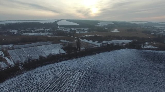 Aerial view of countryside during sunset at winter evening. Plowed fields, village houses, woods among the hills.