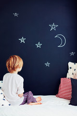 cute kid in pajamas dreaming , while sitting in bed and looking on chalkboard wall
