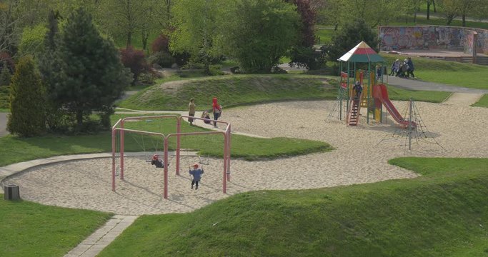 Kids on Playground Landscape Design Trees Lawns Children Swinging on Swings Sandy Ground Parents Standing and Talking Kids Are Playing Sunny Spring Day