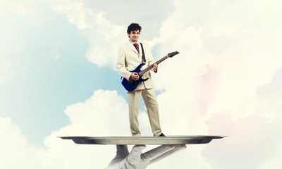 Businessman on metal tray playing electric guitar against blue sky background