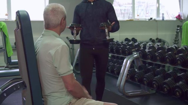 Young caucasian trainer giving dumbbells to the senior client. Old gray man sitting on fitness bench at the gym. Aged male sportsman prepare for exercises