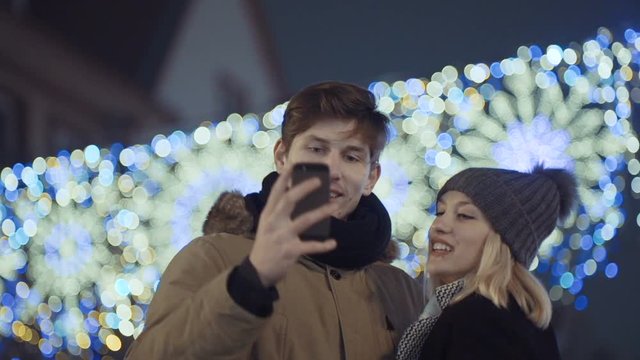 Happy young loving couple making selfie and smiling while standing over Christmas bokeh background. Happy moments together. 