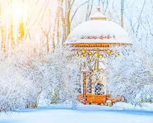 Wooden gazebo pavilion in the winter park forest covered with snow. Sun glowing scene.
