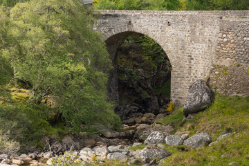 Fototapeta na wymiar Dornoch Firth, Scotland - June 3, 2012: The historic, beige-brown-stone bridge over Allt Fearn Burn, a rocky creek, on the B9176 between Cromarty and Dornoch Firths. Green trees.