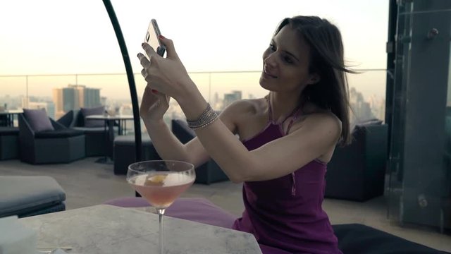 Happy woman with cocktail taking selfie photo with cellphone on terrace in bar 
