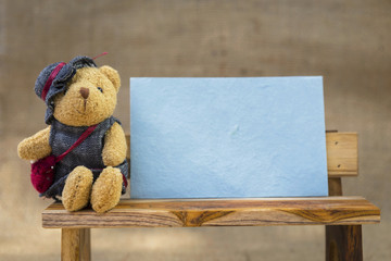 Blank blue paper card and teddy bear on wooden bench over hessian texture background