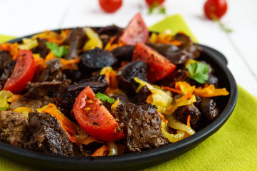 Pieces of roast duck, goose (meat, liver, heart) with vegetables (onion, carrot, tomato), with spices on a round cast-iron frying pan on a green napkin. Close up