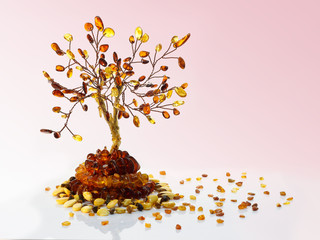 Decorative tree made with yellow and dark brown baltic amber, amber beads and small pieces of raw amber on white acrylic surface on pink gradient background