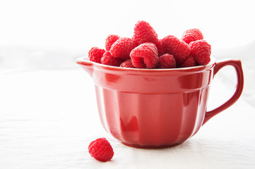 Fresh raspberries in a big red ceramic bowl on a white table.