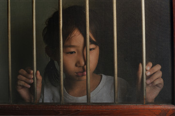 sad asian child standing behind the wire screen window in dark m
