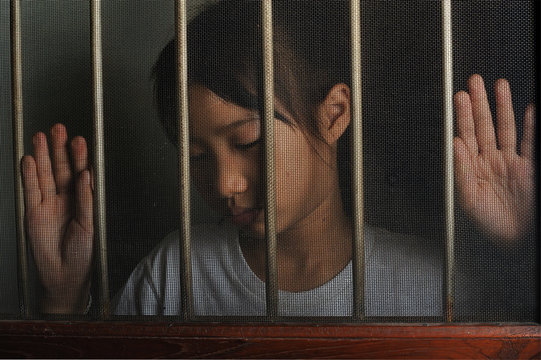 sad asian child standing behind the wire screen window in dark mood.  Unhappy child alone at home. Upset in the dark room