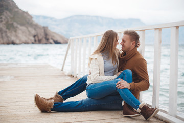 Fototapeta na wymiar Young couple blond guy with short hair in a brown sweater and blue jeans and a blonde girl with straight long hair,spend time together,sitting,embraced on a white wooden pier near the sea in autumn