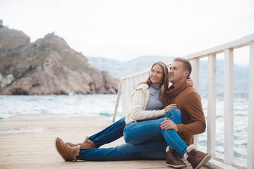 Young couple blond guy with short hair in a brown sweater and blue jeans and a blonde girl with straight long hair,spend time together,sitting,embraced on a white wooden pier near the sea in autumn