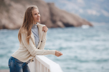 Fototapeta na wymiar Young woman with long straight blond hair and gray eyes, dressed in a white knitted jacket, a gray turtleneck and blue jeans, spending time alone, standing on white wooden wharf near the blue sea 