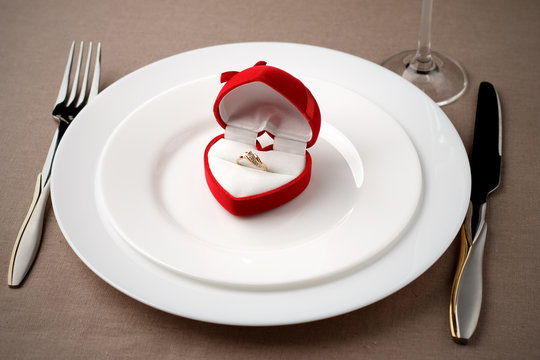 Golden ring in red case on white plate with fork, knife and glass.