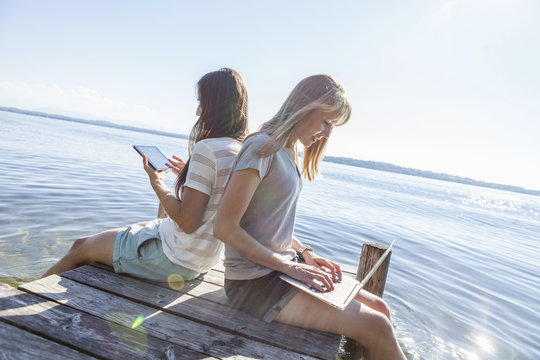 Two young women sit on pier by lake using tablet and laptop