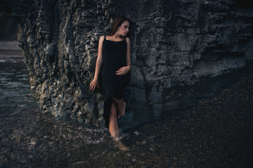 Seductive girl in the black dress standing leaning against the rock