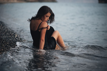 Beautiful girl wearing the black dress lying on the pebbles on the beach in the waves