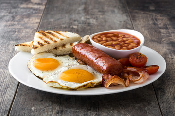 Traditional full English breakfast with fried eggs, sausages, beans, mushrooms, grilled tomatoes and bacon on wooden background
