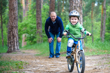 Fototapeta na wymiar Little kid boy of 3 years and his father in autumn forest with a bicycle. Dad teaching son. Man happy about success. Child helmet. Safety, sports, leisure kids concept