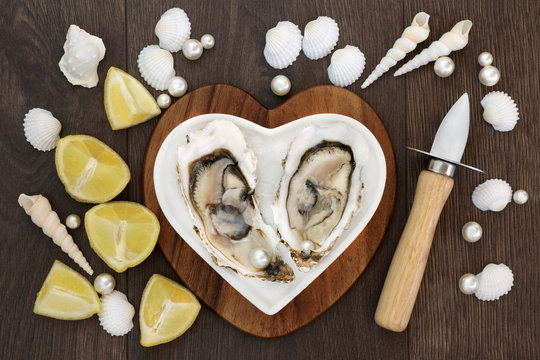 Fruits of the Sea. Oysters on crushed ice on a heart shaped plate on a maple wood board with oyster knife and pearls, lemon fruit and shells over old oak background. Used to stimulate sexual drive.  