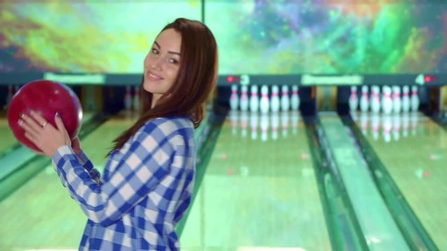 Pretty caucasian girl holding bowling ball in her hands. Attractive brunette lady in checkered shirt smiling for the camera. Beautiful female bowler posing against background of lanes
