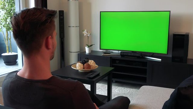 A man watches a TV with a green screen in a cozy living room