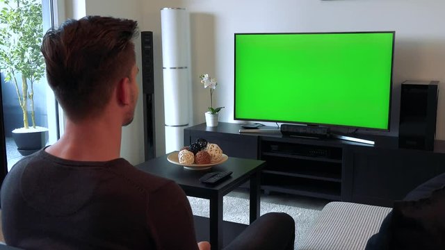 A young, handsome man watches a TV with a green screen, then turns to the camera and smiles