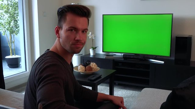 A young, handsome man watches a TV with a green screen, then turns to the camera and shakes his head, unhappy