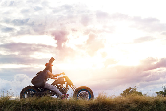 Guitarist woman riding a motorcycle on the countryside road, sunset background