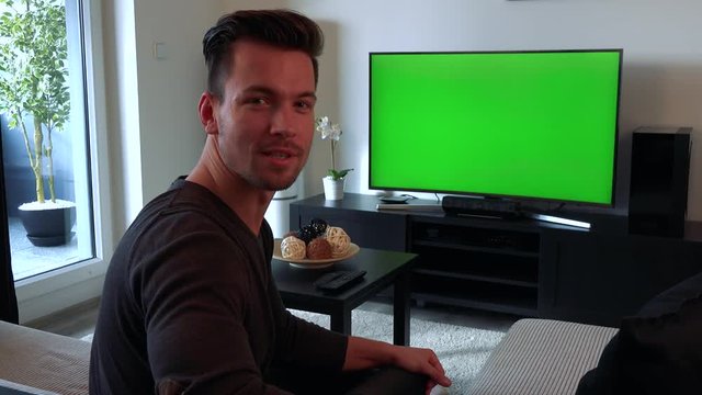 A young, handsome man watches a TV with a green screen, then turns to the camera and talks to you with a smile as if explaining something about the TV
