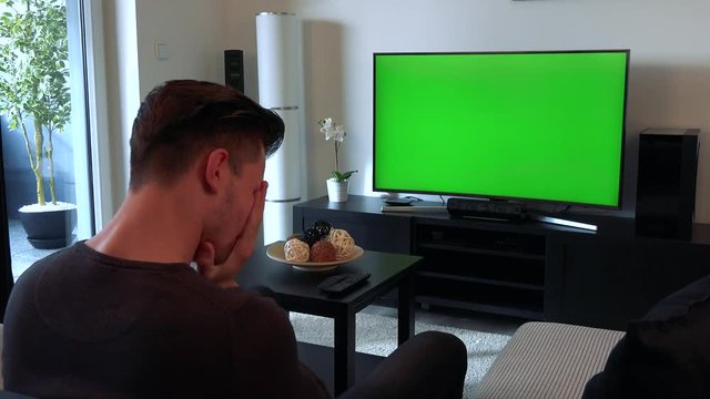 A man watches a TV with a green screen in a cozy living room and then buries his face in his arms, upset and disappointed