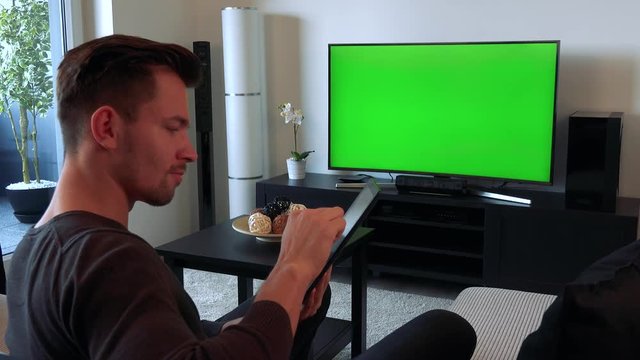 A young, handsome man watches a TV with a green screen in a cozy living room and types on his tablet