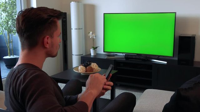 A young, handsome man watches a TV with a green screen in a cozy living room and types on his smartphone