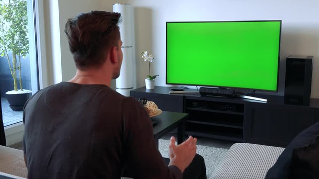 A man cheers and gesticulates excitedly at a TV with a green screen