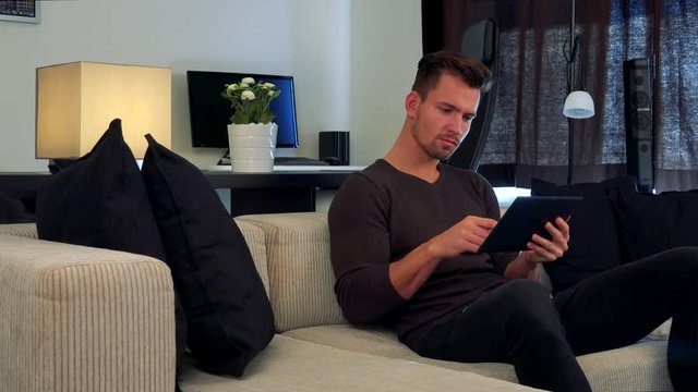 A young, handsome man sits on a couch in a cozy living room and uses his tablet