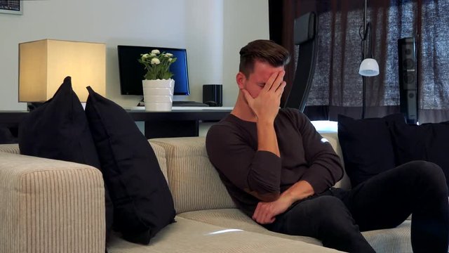 A young, handsome man on a couch covers his face with a palm, disappointed