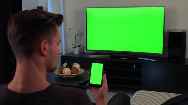 A man turned with the back of his head to the camera watches a TV and eventually glances at a smartphone in his hand, both the TV and the phone have a green screen