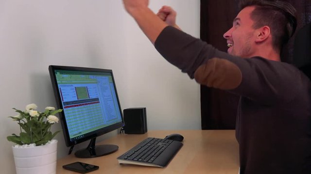 A young, handsome man sits at a computer and celebrates