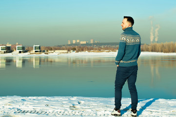 man in warm sweater with a beard is walking on the street in the winter in a warm sunny day at the river against the backdrop of the city