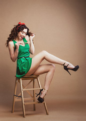 Obraz na płótnie Canvas Curly Pin-up girl in green dress sitting on chair, having fun and her perfect legs at rough paper background. Stylish studio shot for poster.