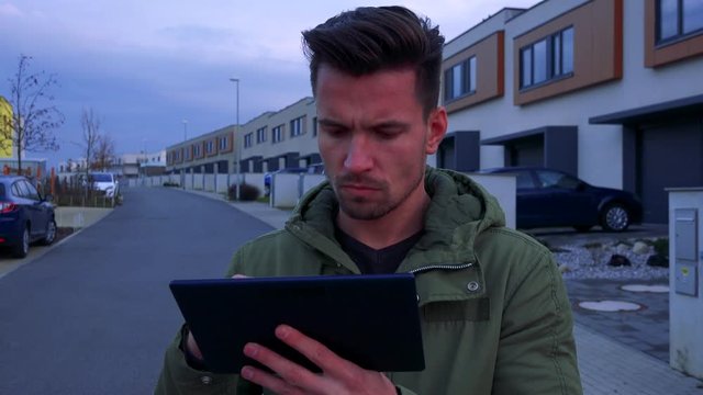 A young, handsome man stands on a road in a neighborhood and works on a tablet