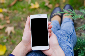 Close up white mobile phone in hand a young hipster business woman in denim blue jeans pants on the background of green natural grass and autumn leaves