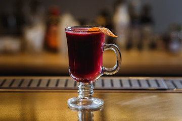 A transparent cup of raspberry mulled wine with orange peel on a bar stand. Selected focus, small depth of field, film grain effect. 