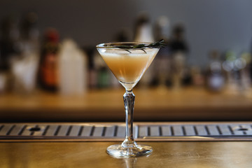 A drink on the bar stand in a cocktail (martini) glass with rosemary on top. Small depth of field, selective focus, film grain effect.