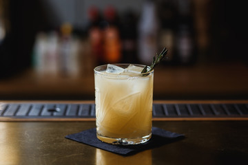 A cocktail on the bar stand in a whisky glass, with ice cubes inside and rosemary.  Selective focus, small depth of field, film grain effect.