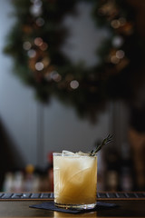 A cocktail on the bar stand in a whisky glass, with ice cubes inside and rosemary. Christmas wreath in the background. Selective focus, small depth of field, film grain effect.